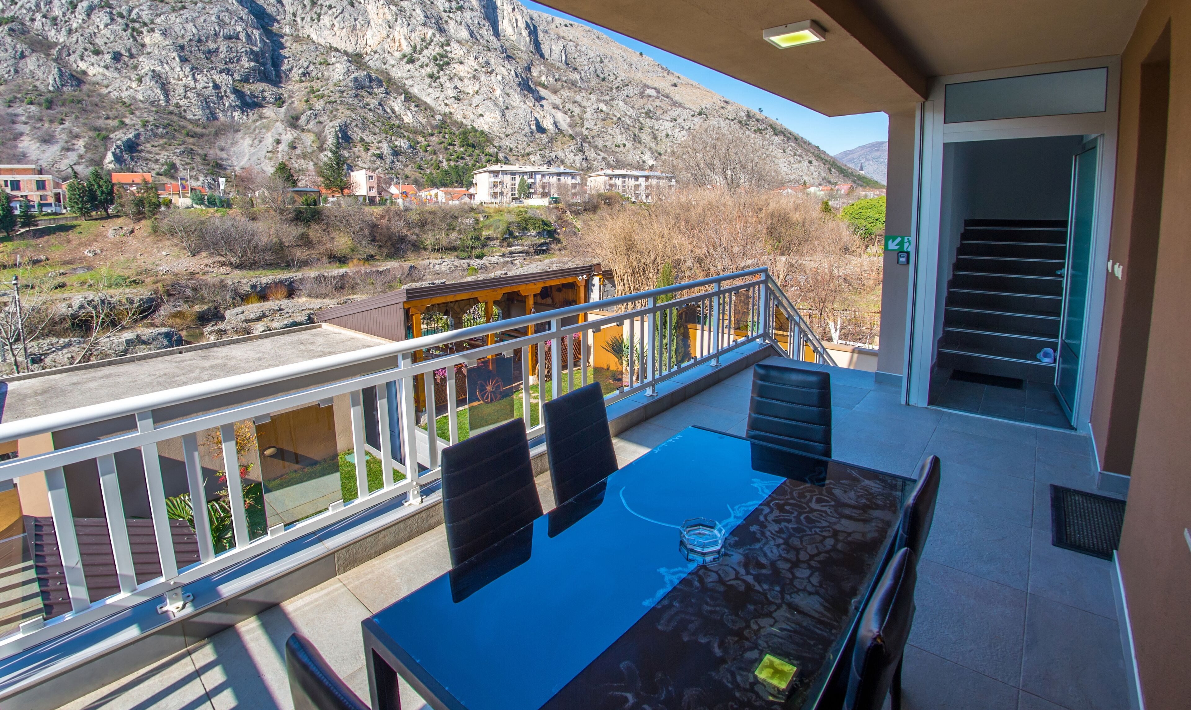 Un balcon avec une table et des chaises, donnant sur un paysage montagneux avec des bâtiments visibles au loin.