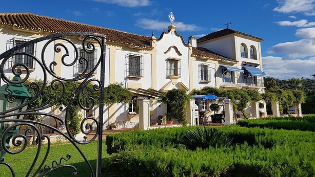 Un edificio grande y histórico con fachada blanca y techo de tejas rojas, rodeado de jardines bien cuidados y una valla de hierro decorativa.