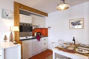 Cocina compacta con armarios blancos, estufa y comedor con mesa para dos.
