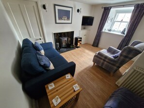 Ein Wohnzimmer mit einer Couch, einem Sessel, einem kleinen Tisch, einem Kamin und einem an der Wand montierten Fernseher.