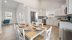 A modern kitchen with white cabinetry, a stainless steel refrigerator, and a dining area with a wooden table set for four. - Stunning Home with Expansive Pool & Game Room (Kemah Area)
