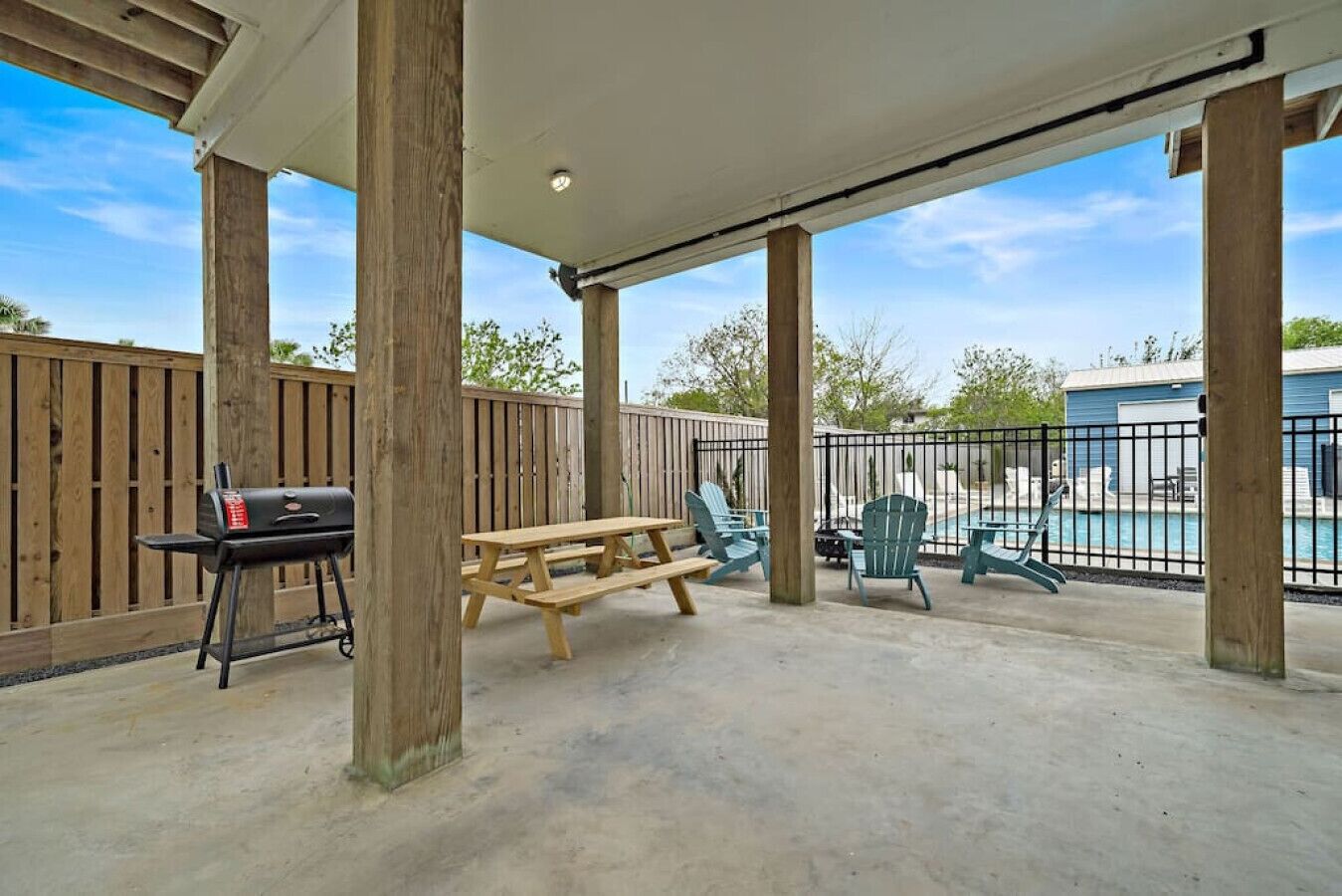 A covered patio area with a picnic table, chairs, a grill, and a pool in the background.