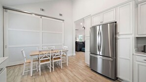 A modern kitchen with white cabinetry, a stainless steel refrigerator, and a dining area with a wooden table and white chairs. - Stunning Home with Expansive Pool & Game Room (Kemah Area)