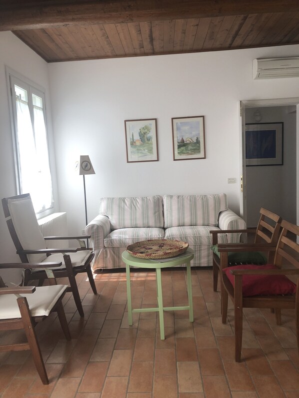 A living room with a striped sofa, a wooden chair, a small table, and a radiator. There are two framed pictures on the wall and a window with curtains. - Cascina delle Rondini Charme Country House (Sant'Alberto)