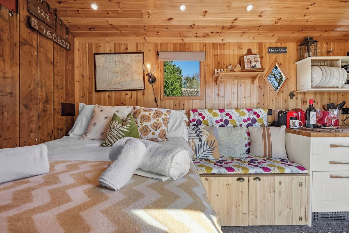A wooden cabin bedroom with a bed, a bench, a map on the wall, and a shelf with dishes.