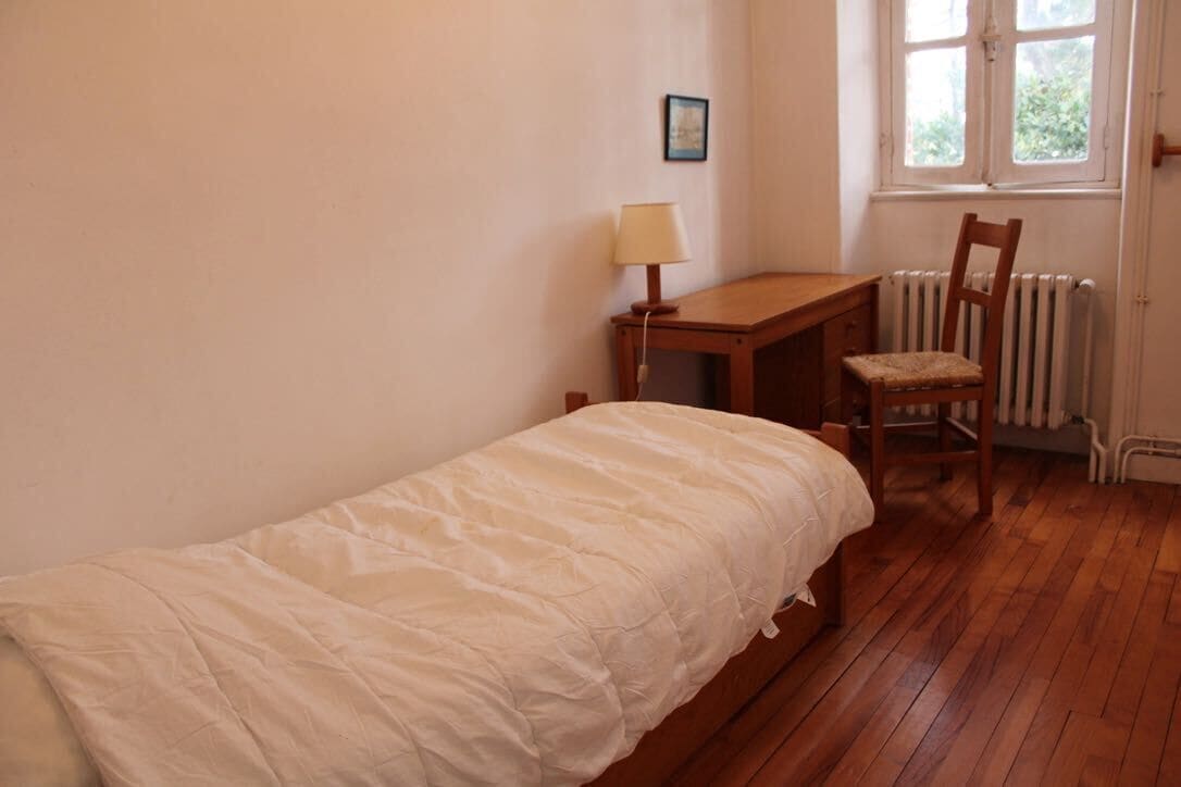 Ein kleiner, ordentlicher Raum mit einem Einzelbett, einem Holztisch, einem Stuhl, einer Heizung und einem Fenster.