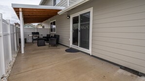 Un patio techado con una parrilla y zona de estar, una puerta corrediza de vidrio y una cerca blanca.