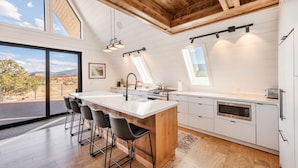 A modern kitchen with a large island, wooden ceiling beams, and large windows offering a view of the outdoors.