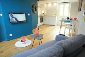 Un salon moderne avec un canapé gris, une petite table blanche surmontée d’un vase en verre rose et un téléviseur fixé au mur.