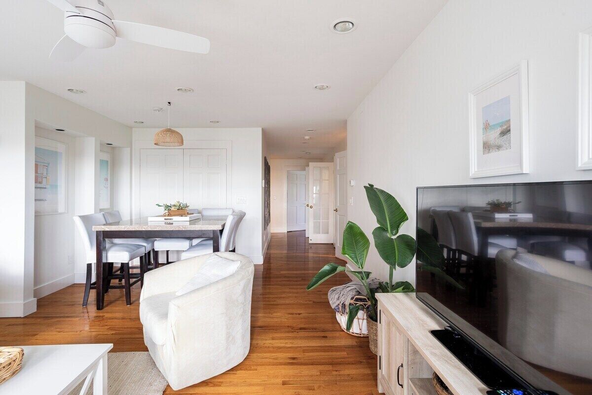 A modern living room with a flat-screen TV, a white sofa, a dining area with a table and chairs, and a large plant.