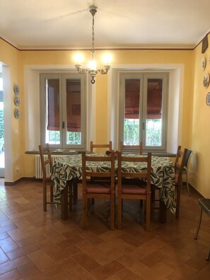Un comedor con una mesa dispuesta para cuatro personas, sillas de madera, una araña y ventanas con persianas.