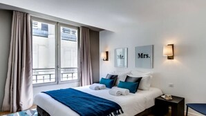 A hotel room with a bed, a nightstand, a chair, and a window with curtains. - A Bit of Glamour and Magic in Champs Elysees (Paris)