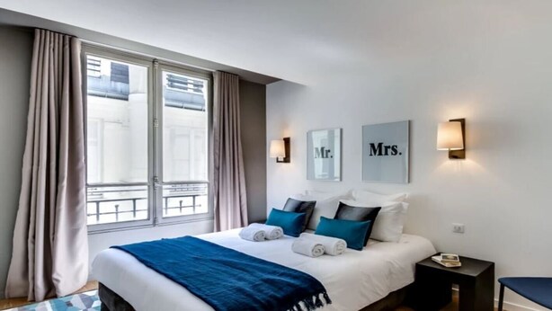 A hotel room with a bed, a nightstand, a chair, and a window with curtains. - A Bit of Glamour and Magic in Champs Elysees (Paris)