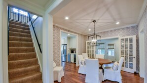 A spacious living area with a wooden staircase, a dining table with chairs, and a kitchen in the background.