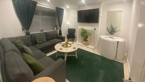 A modern living room with a grey sofa, a small round table, a flat-screen TV mounted on the wall, and a fireplace. - Charming 3-bed House Near Central Manchester (Manchester)