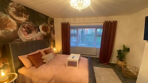 A bedroom with a large bed, a bedside table, a nightstand, a window with curtains, a chandelier, and a potted plant. - Charming 3-bed House Near Central Manchester (Manchester)