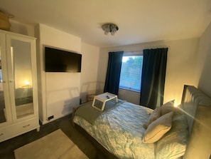 Ett sovrum med en säng, ett sängbord, en tv som är monterad på väggen, en garderob och ett fönster med gardiner.