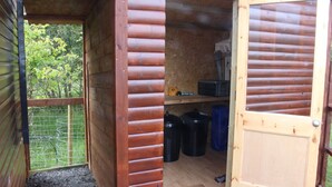 A wooden cabin with a door open, revealing a small interior with a shelf, black trash bins, and a blue bag.