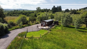 A small wooden cabin with a porch and a gravel driveway, surrounded by lush greenery and trees.