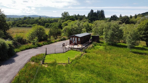 A small wooden cabin with a porch and a gravel driveway, surrounded by lush greenery and trees. - Loch View - luxury cabin with spectacular alpine and loch views (Taynuilt)