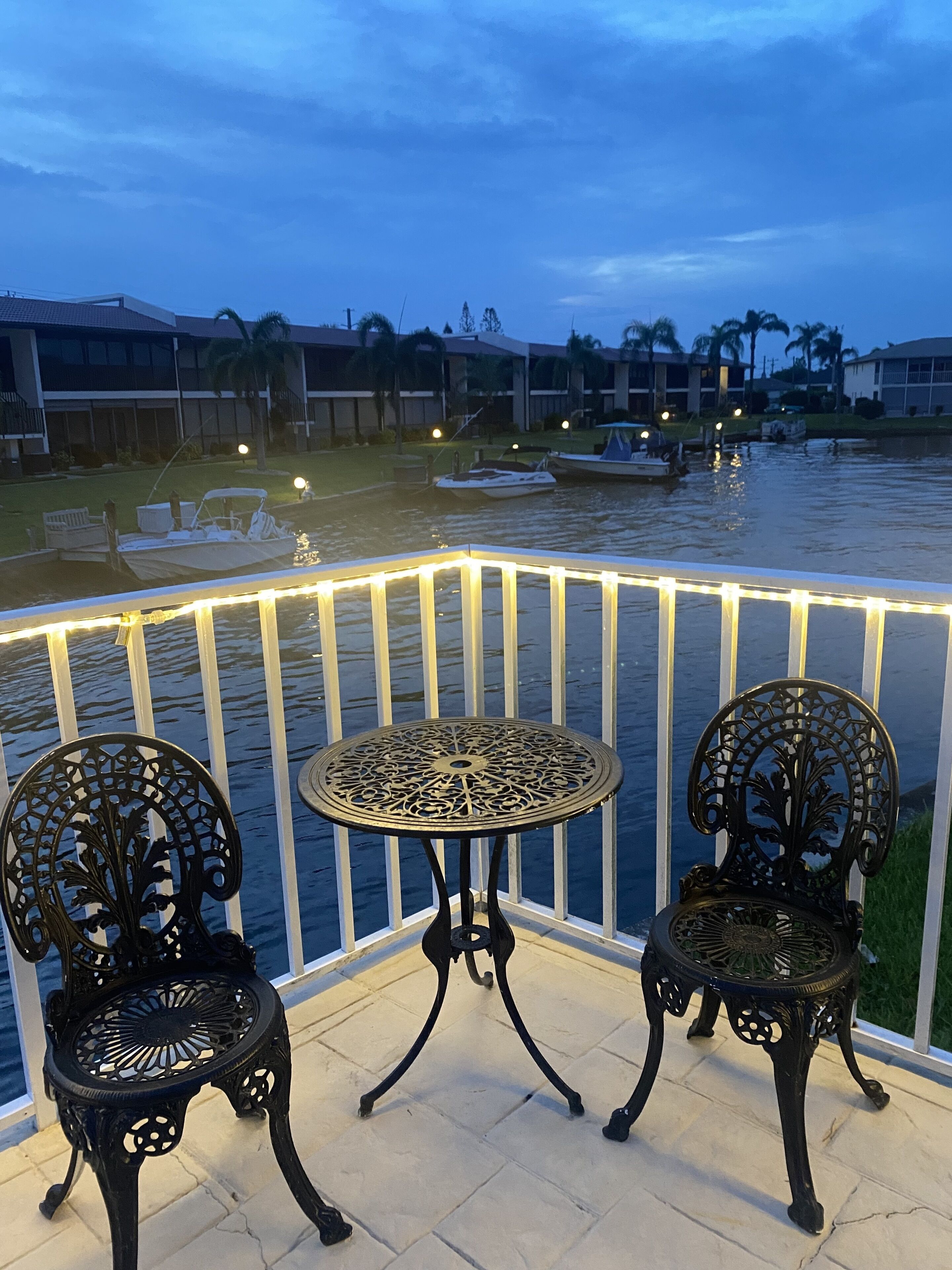 A balcony with two ornate metal chairs and a small table, overlooking a body of water with boats and a building in the background.