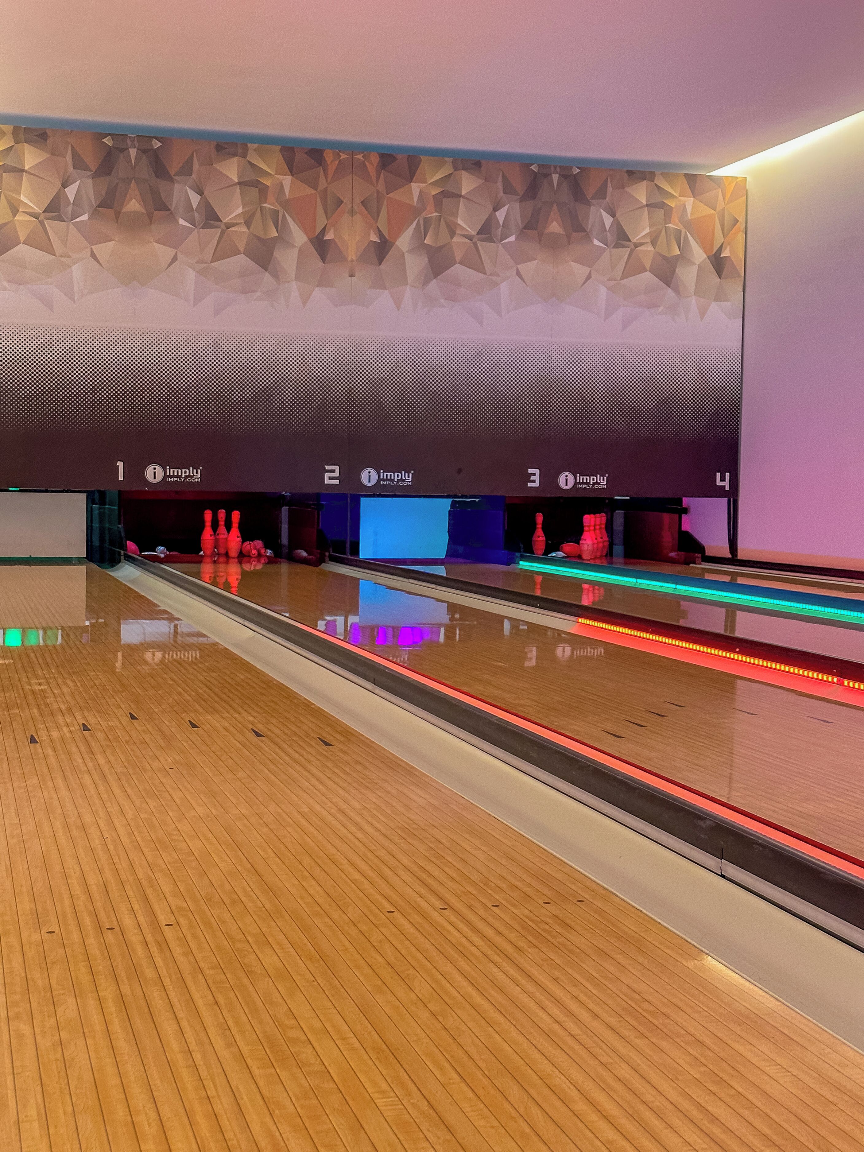 A bowling alley with multiple lanes, wooden flooring, and a modern wall with geometric patterns.
