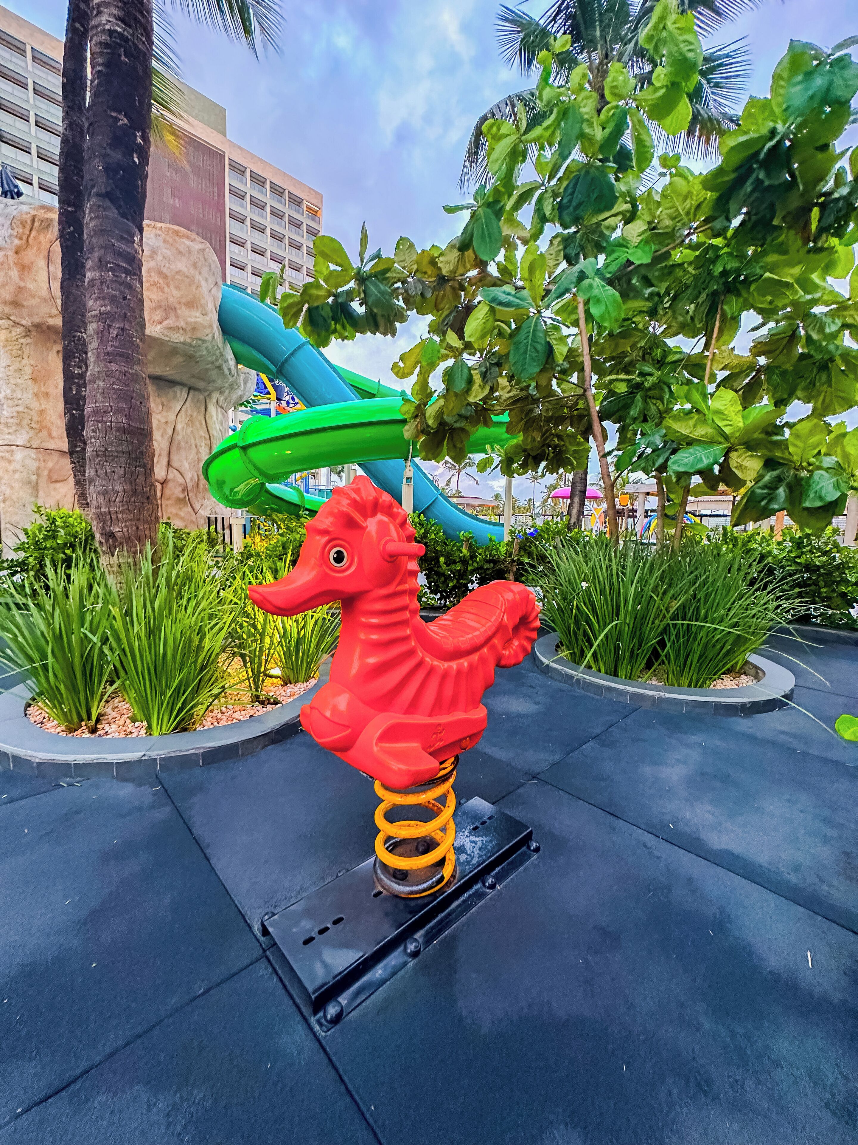 Una escultura de caballito de mar rojo sobre una plataforma negra en un área ajardinada, con palmeras y un tobogán azul al fondo.