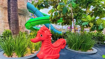 Una escultura de caballito de mar rojo sobre una plataforma negra en un área ajardinada, con palmeras y un tobogán azul al fondo.