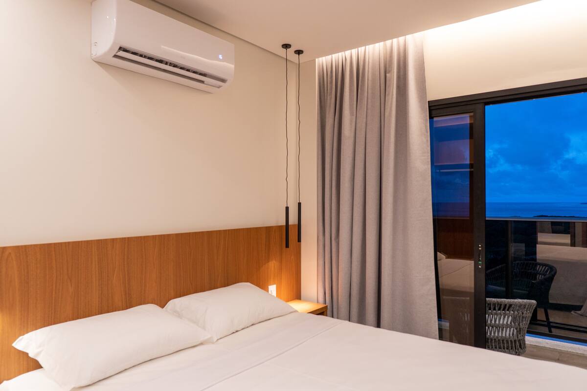 A hotel room with a bed, an air conditioning unit, a nightstand, a chair, and a view of the ocean through a glass door.