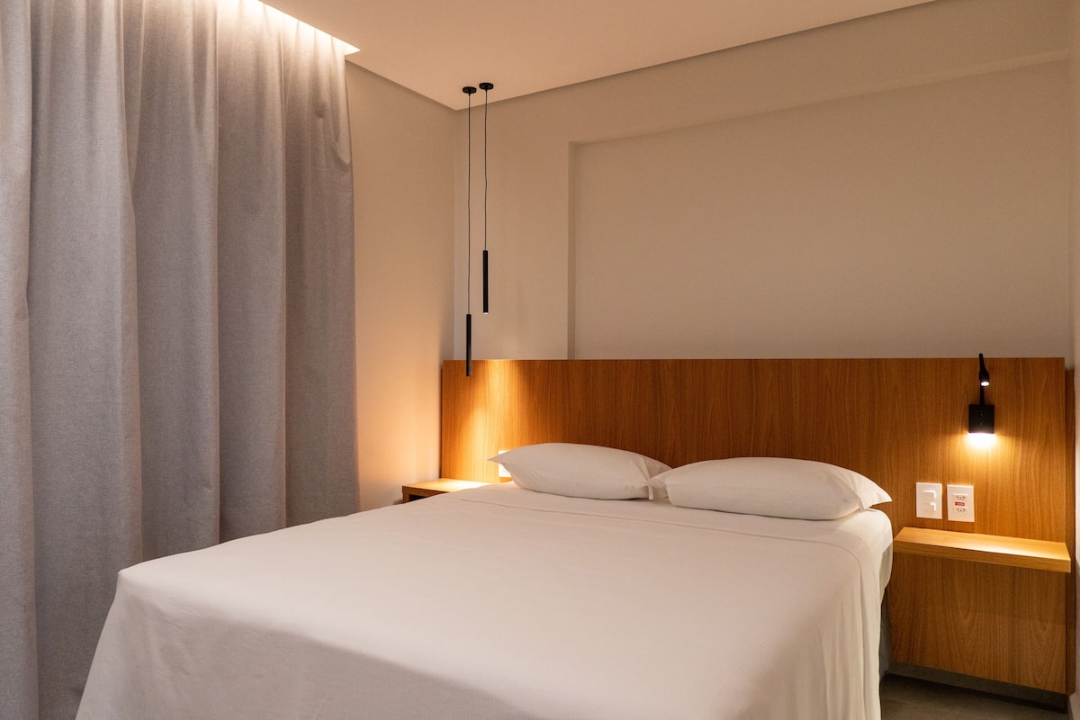 A modern hotel room with a neatly made bed, wooden headboard, bedside tables with lamps, and a large window with curtains.
