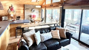 A cozy living room with a black leather sofa, a wooden coffee table, and a kitchen area in the background. - “The Lake house” Private BEACH; SLEEPS 16!!! (Schroon Lake)