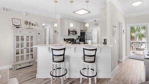 A modern kitchen with white cabinetry, a central island with bar stools, pendant lighting, and a view to the outdoors. - Snowbird Special Jan Feb! 🌊Canal View~pools~Walk2Beach~2BR Sandpiper Cove 4114 (Destin)
