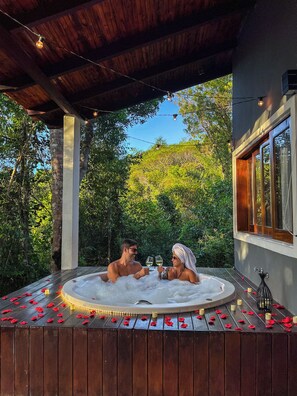 A couple in a hot tub with rose petals and candles, surrounded by greenery and a wooden deck. - Chalés Pura Vida (Itacaré)