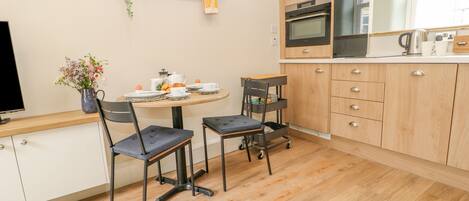 Een moderne keuken met houten kastjes, een inbouwoven en een koffiezetapparaat. Er is een eettafel met stoelen en een kleine plant aan de muur.