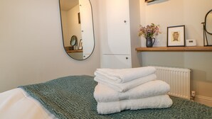 Un lit bien fait, recouvert d’une couverture verte, une pile de serviettes blanches, un miroir, un radiateur et une étagère ornée d’objets décoratifs.