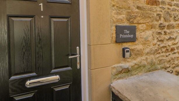 Une porte en bois sombre, dotée d’un clavier numérique et d’une petite fenêtre ornée d’un motif géométrique. Un mur de pierre sur lequel est fixée une plaque portant l’inscription « The Printshop » est visible sur la droite.