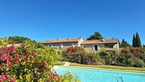 A poolside view of a villa with a clear blue sky, surrounded by lush greenery and flowering plants. - Alpilles Cottage Philippe's Farm pool shared (Saint-Rémy-de-Provence)