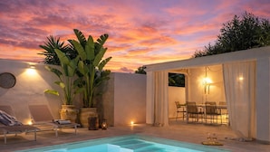 Un espace piscine extérieur moderne offrant une vue sur le coucher du soleil, avec des chaises longues et un coin repas aménagé.