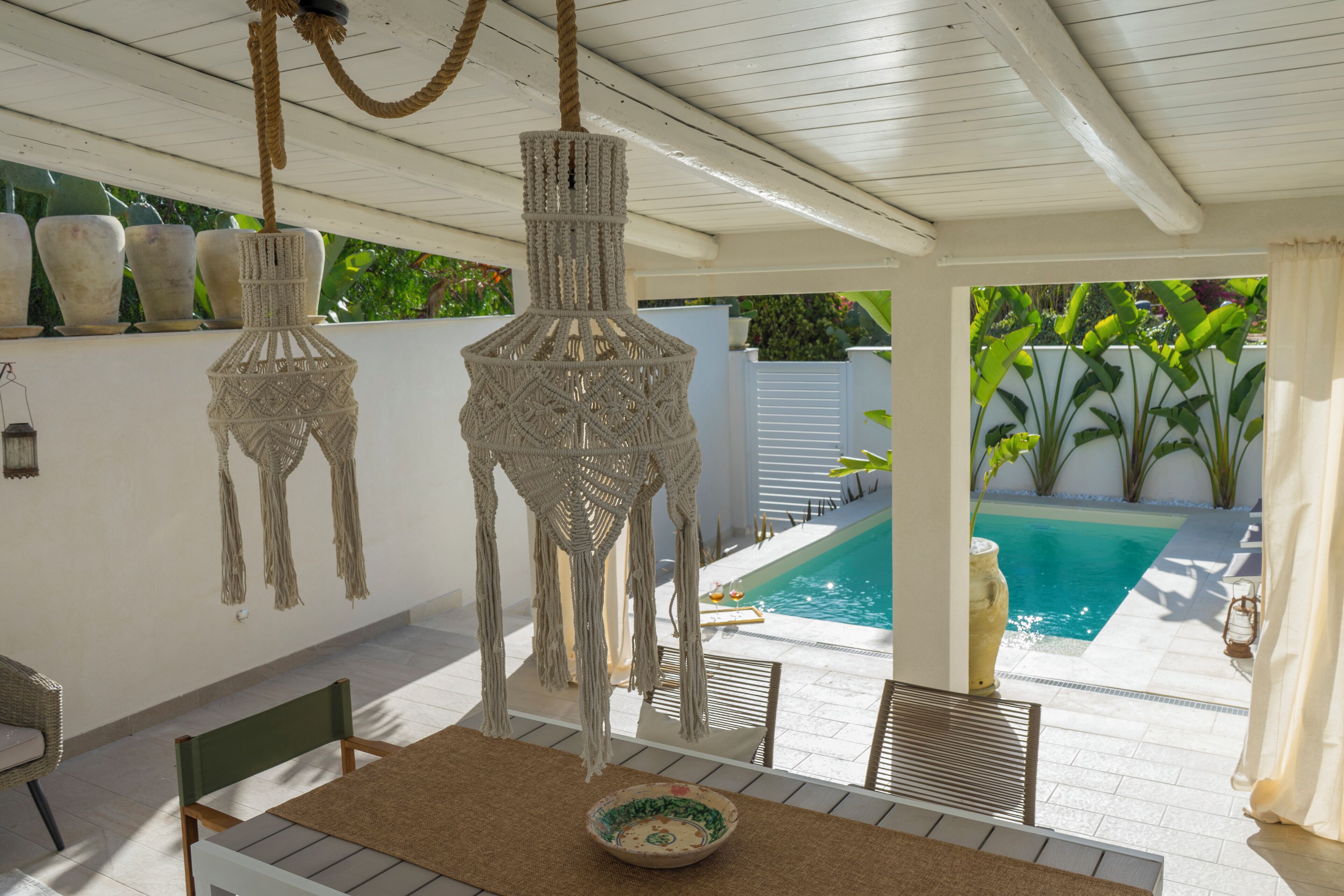 Zona junto a la piscina con una mesa, sillas, lámparas de macramé y vistas a una piscina.