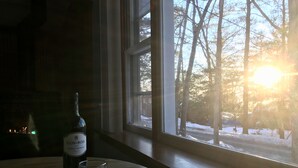 A bottle of Clos du Bois wine and a glass on a wooden table by a window with a view of trees and a snowy landscape. - Cozy Lakeview Family Cottage - steps to the lake, 2 swim docks & restaurant! (Hawley)