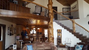 A spacious cabin interior with a high ceiling, wooden beams, a dining area, and a staircase.