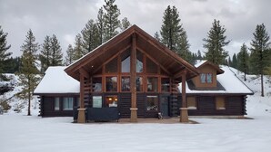 A log cabin with a large porch and multiple windows, surrounded by snow-covered trees.