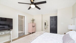 Un dormitorio con cama, ventilador de techo, televisor, cómoda, puerta y lámpara.