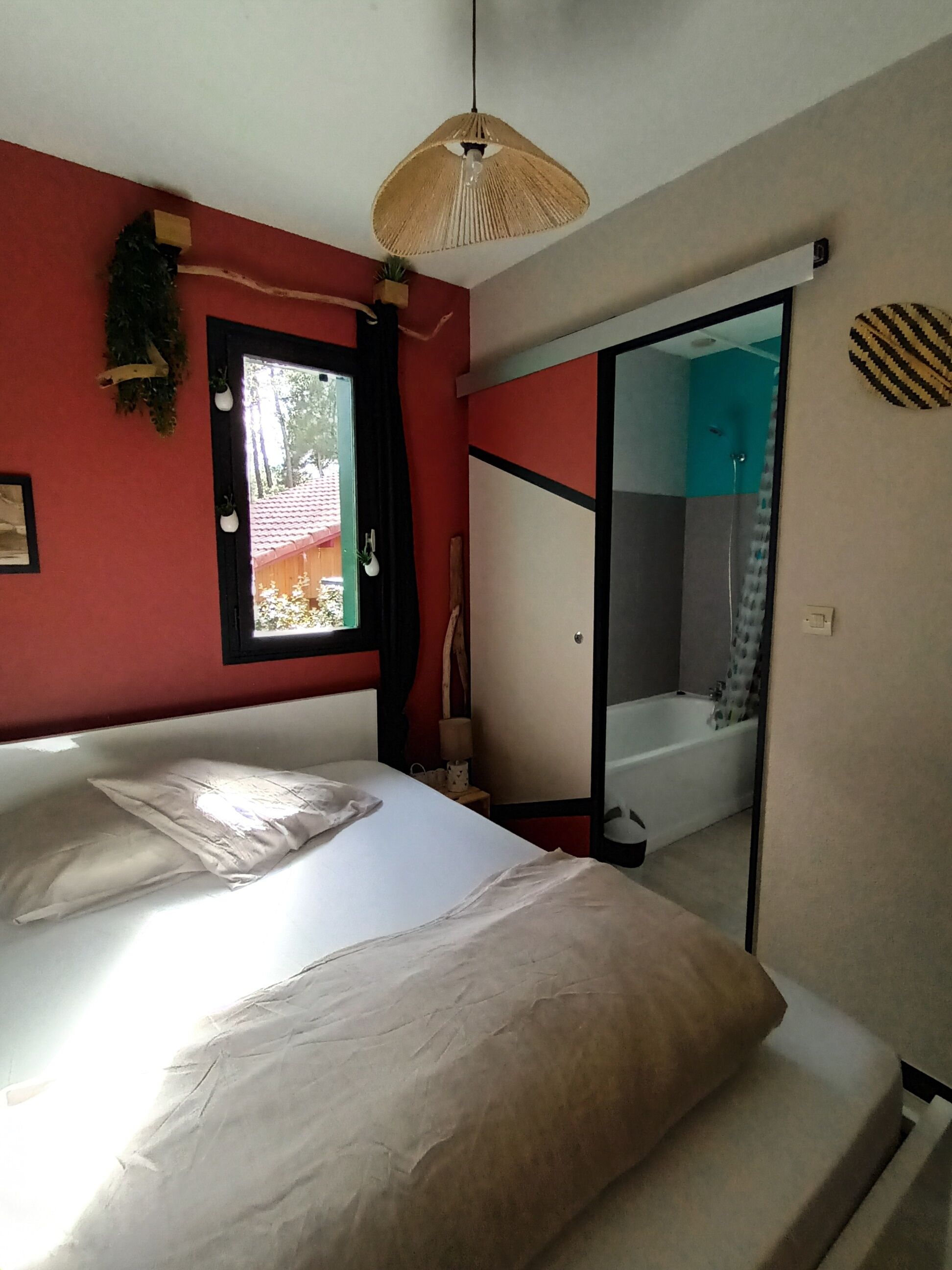 A bedroom with a bed, a hanging light fixture, a window, and a bathroom visible through a sliding door.
