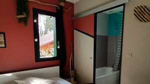 Une chambre à coucher avec un lit, un luminaire suspendu, une fenêtre et une salle de bain visible à travers une porte coulissante.