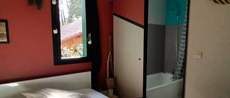 A bedroom with a bed, a hanging light fixture, a window, and a bathroom visible through a sliding door.