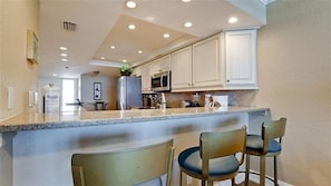 A kitchen with white cabinets, a granite countertop, and a stainless steel refrigerator. There are two bar stools and a microwave on the counter. - Anna Maria Island Club 11, Condo 2 Bedroom/ 2 Bath Gulf view, maximum occupancy of 4 people. Community pool, Max 1 vehicle. (Bradenton Beach)