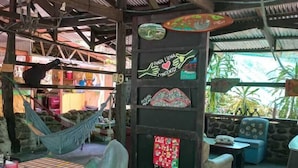 Un área de estar al aire libre rústica con una hamaca, una mesa con mantel a cuadros, y una pared con arte colorido y un cartel de Coca-Cola.