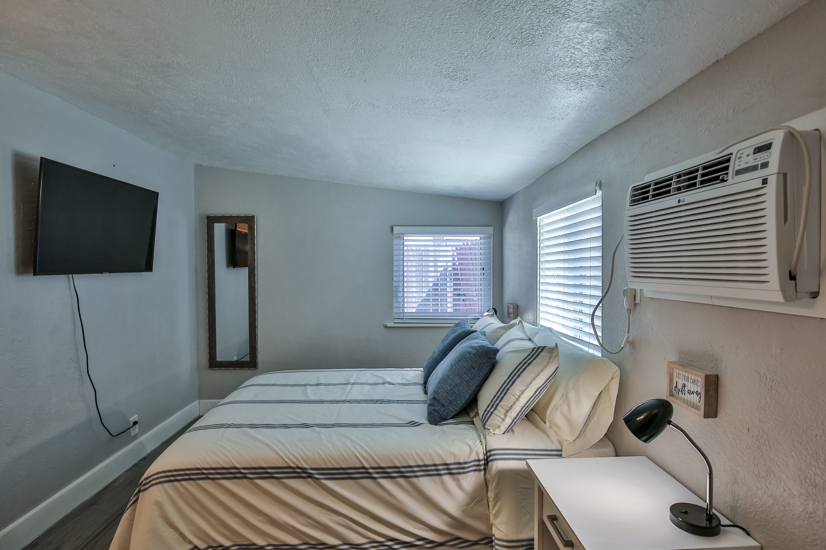 A bedroom with a bed, a TV mounted on the wall, a window with blinds, an air conditioner, and a bedside table with a lamp.