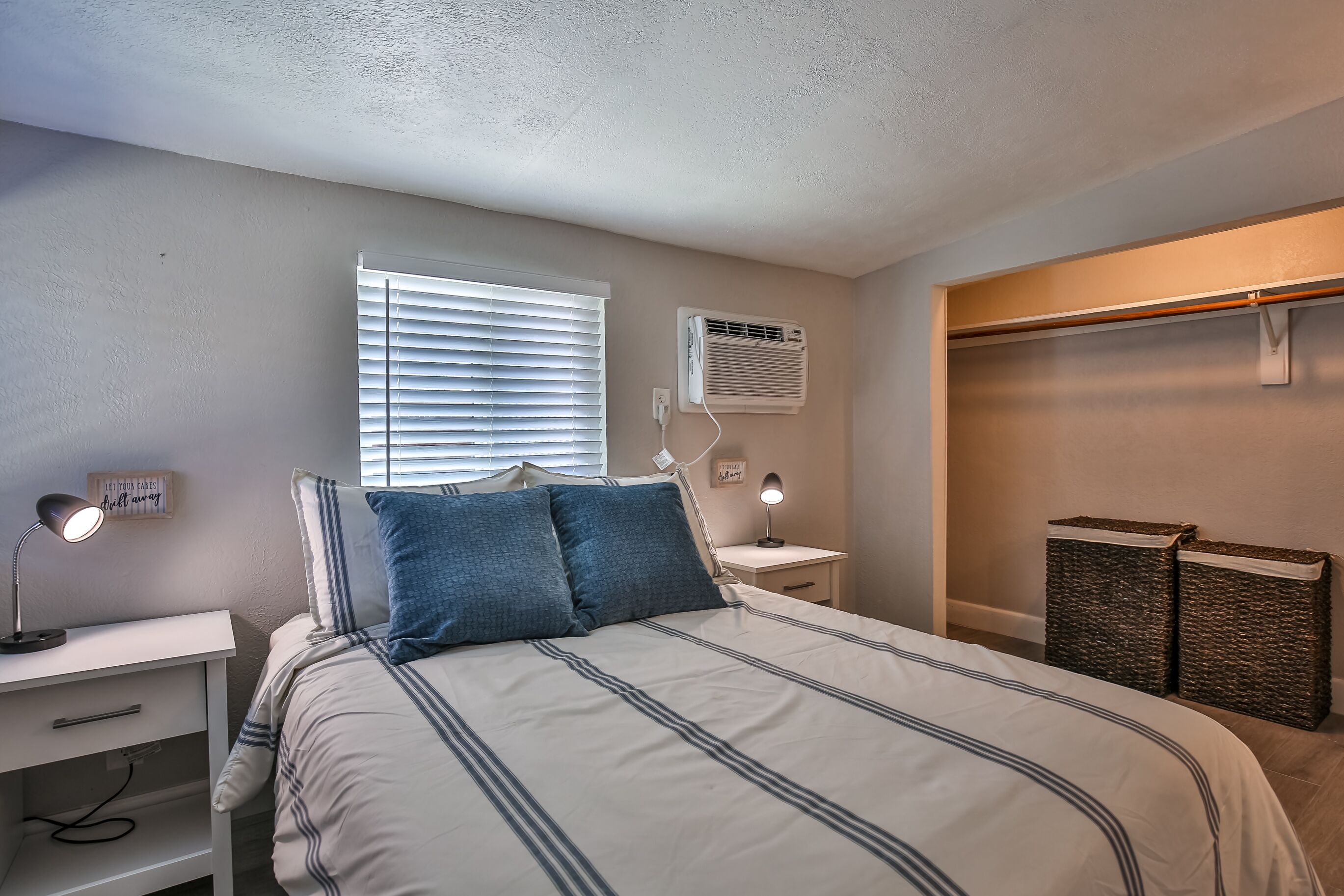 Une chambre à coucher comprenant un lit, des tables de chevet, une lampe, un climatiseur et une fenêtre avec des stores.